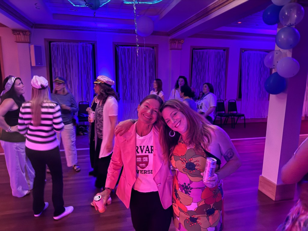 Two women smiling together at a party, with colorful decorations and guests in the background. The atmosphere is lively with purple lighting.