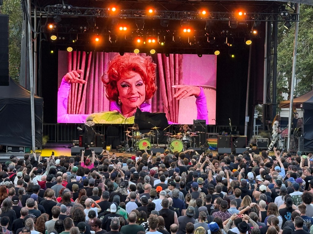 A lively outdoor concert scene featuring a large crowd watching a performance on stage, with a colorful big screen displaying a smiling woman with red hair in vibrant attire.