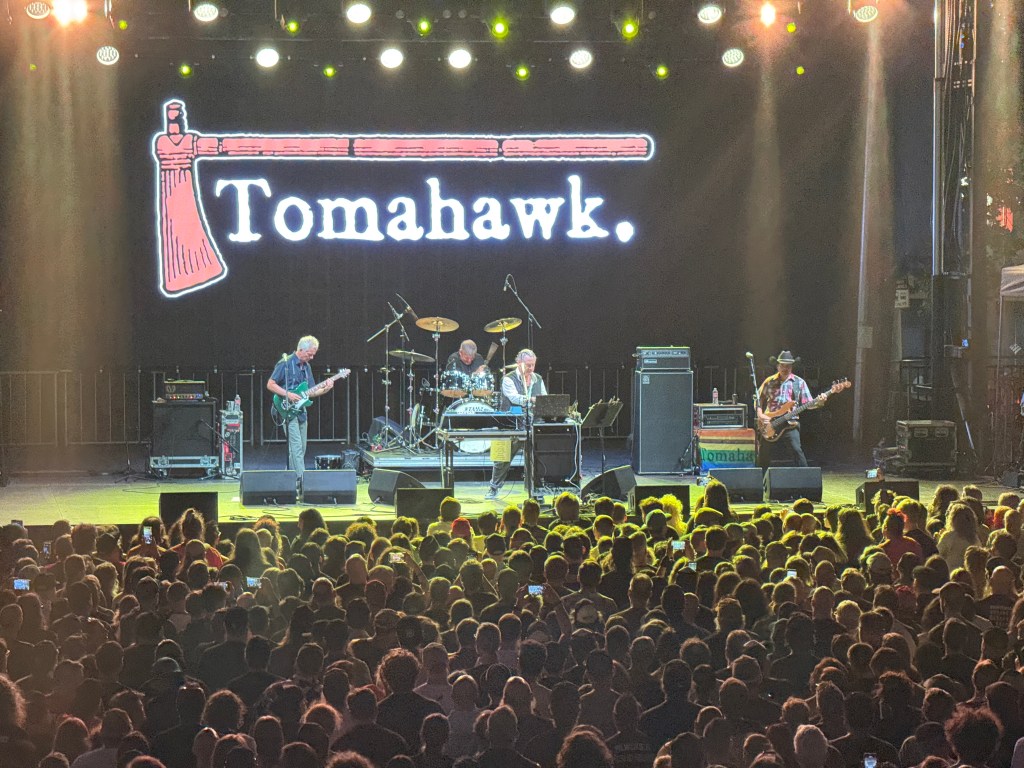 A live music performance by the band Tomahawk on stage, with a large audience in front. The stage features lighting effects and a backdrop displaying the band's name.