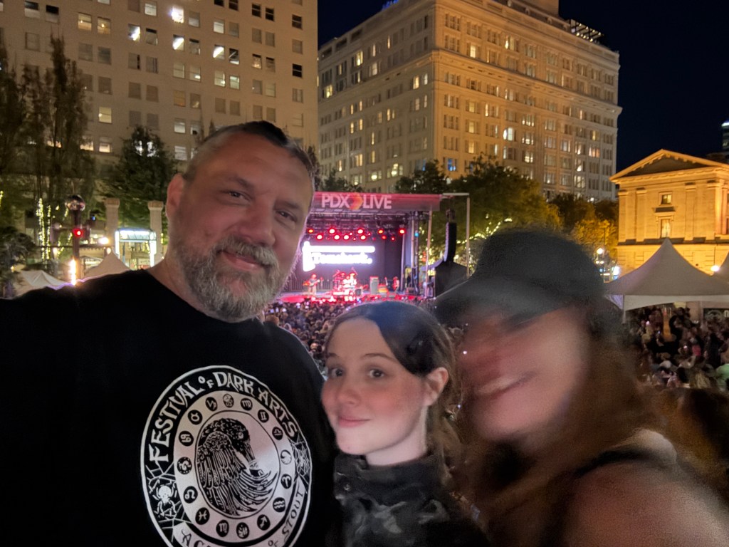 A group of three people at a live concert, smiling for the camera with a stage and audience visible in the background under night lighting.
