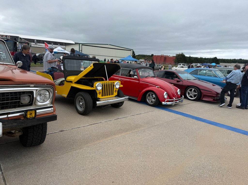 A row of classic cars displayed at a car show, including a yellow jeep with its hood open, a red convertible Volkswagen Beetle, and a maroon sports car, with people gathered around.