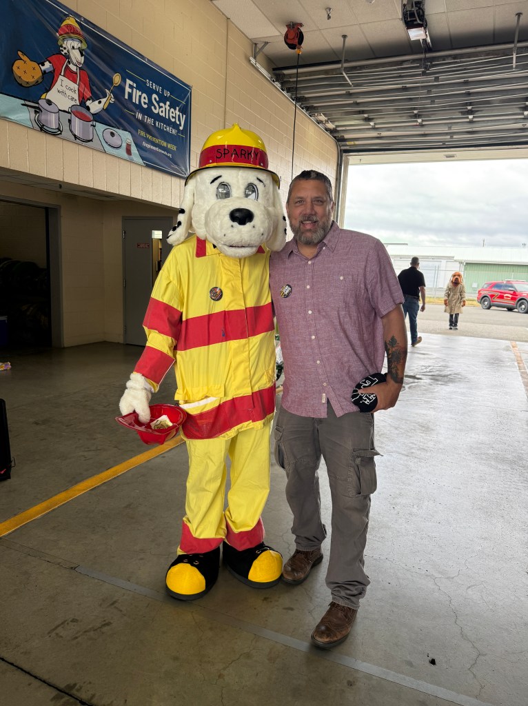 A man poses with a fire safety mascot, Sparky, in a fire station. The mascot is dressed in a firefighter uniform, and the background features a fire safety banner.