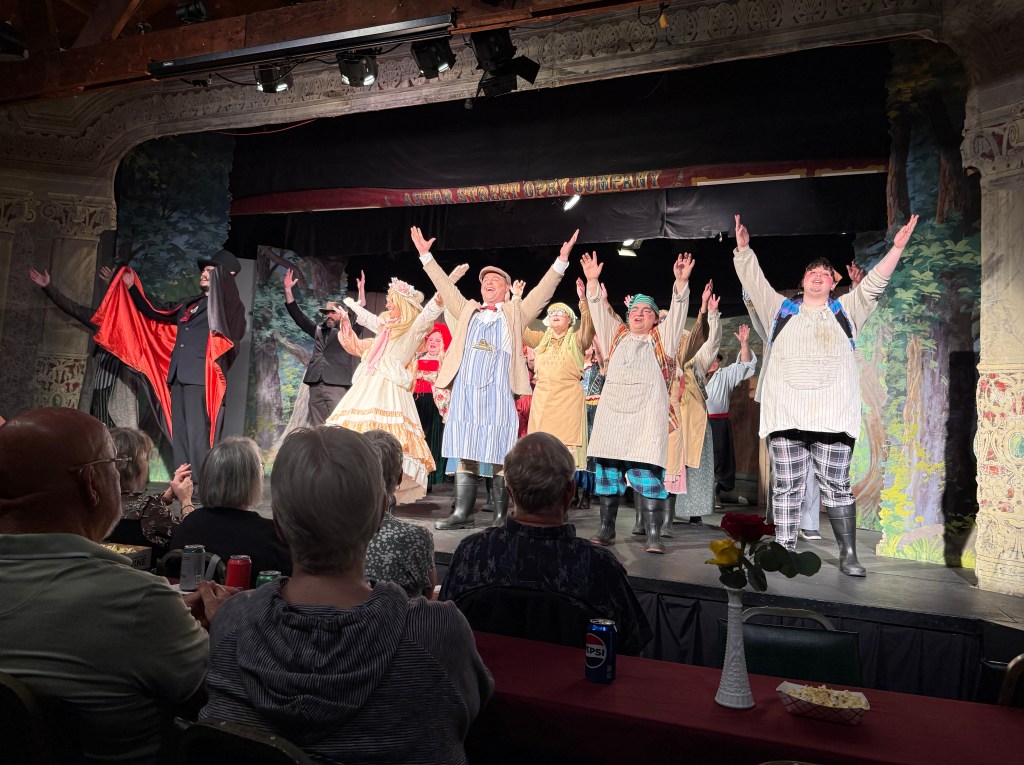 A theatrical performance featuring a cast of actors in costumes, joyfully singing and dancing on stage with their arms raised. The audience is visible in the foreground, enjoying the show.