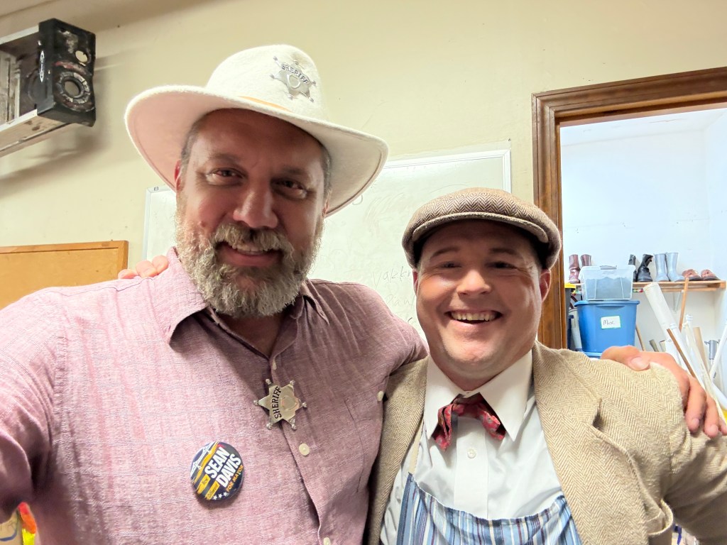 Two men posing for a selfie, wearing costumes; one in a sheriff's outfit with a badge and the other in vintage attire with a bowtie and flat cap.