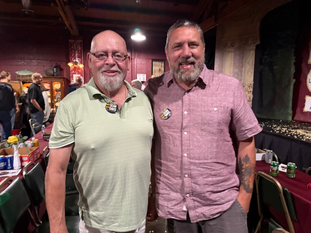 Two men posing for a photo indoors, wearing badges and smiling, with a festive background and tables set for a gathering.