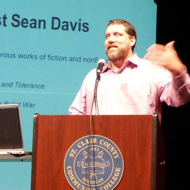 A speaker presenting at a podium with a computer next to him, discussing various works of fiction and nonfiction.