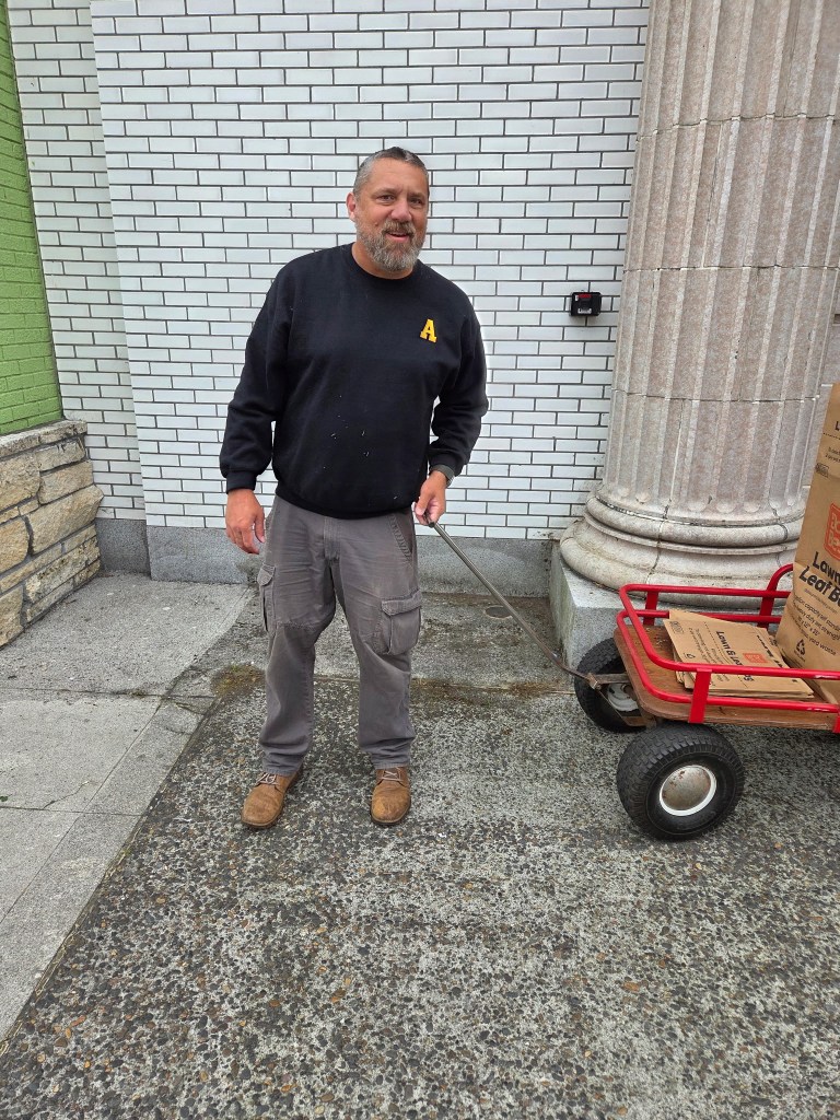A man standing on a sidewalk next to a handcart, smiling at the camera. He wears a black sweatshirt with an 'A' on it and gray cargo pants.