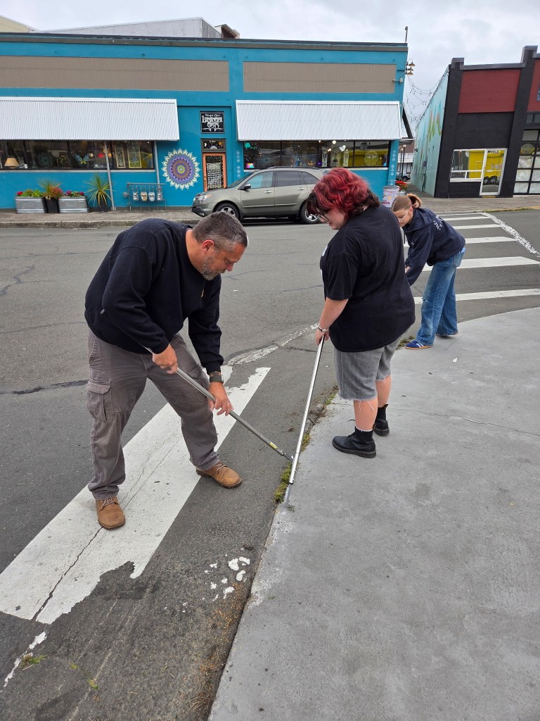 Two people cleaning the gutter in the street with long-handled tools, while a third person observes. A colorful building is in the background.