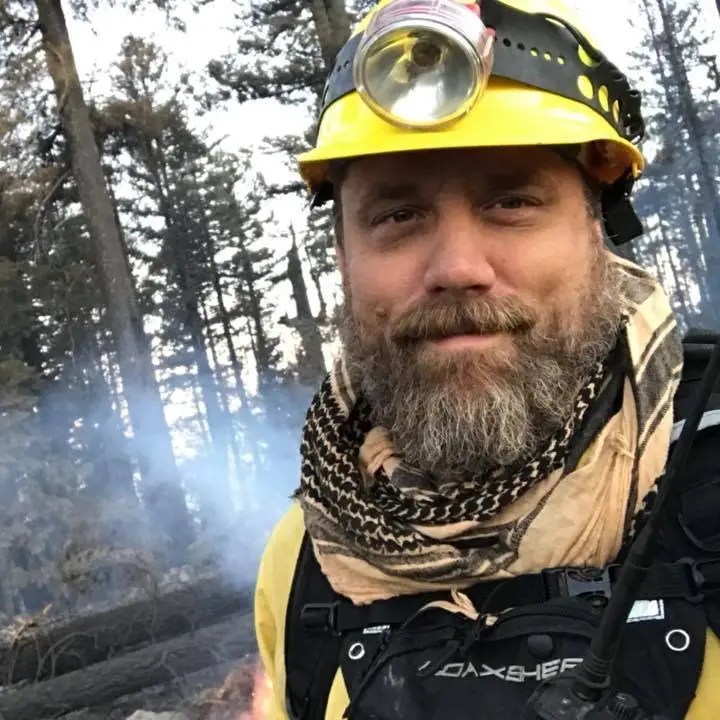 A firefighter with a yellow helmet and headlamp, wearing a black vest and a patterned scarf, stands in a forest with smoke in the background.