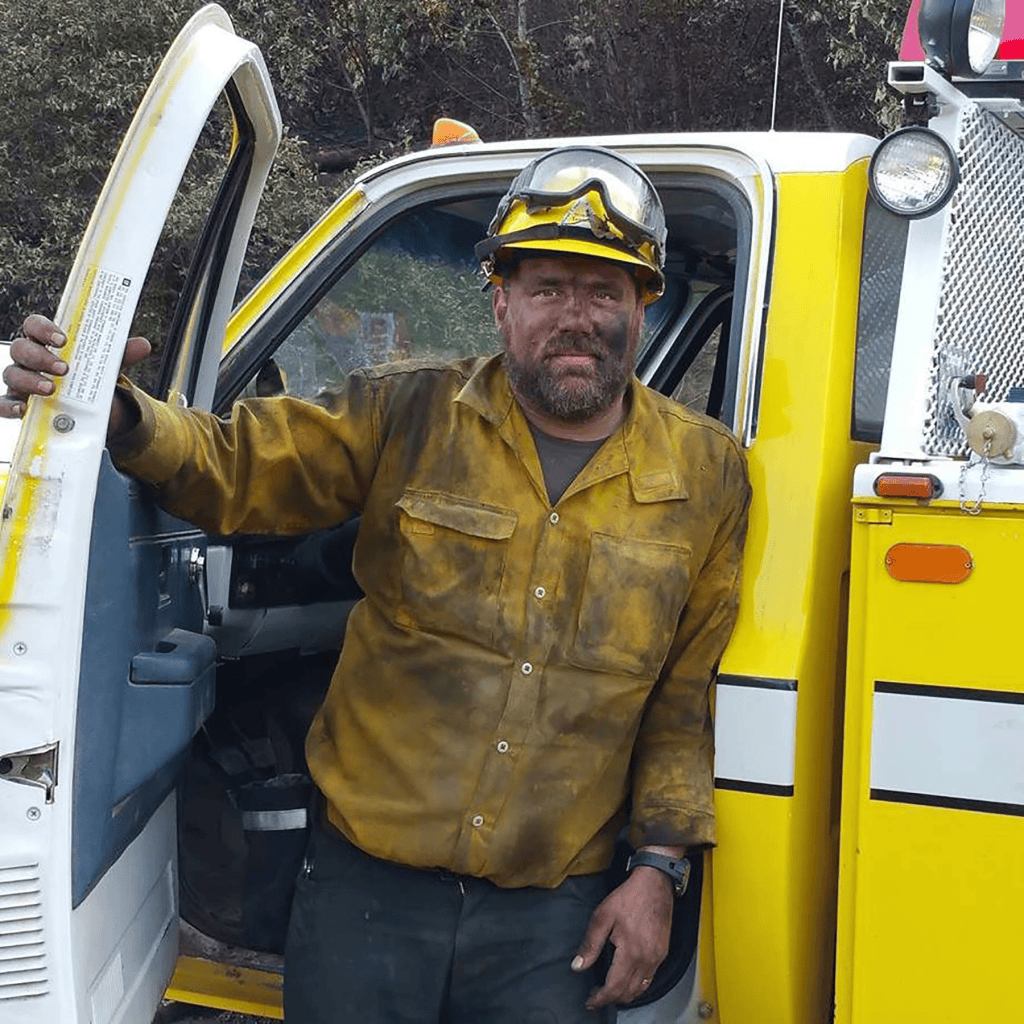 A worker in a yellow shirt and helmet stands next to a yellow vehicle, covered in dirt and grime, indicating his recent labor. He has a serious expression and appears to be taking a break.