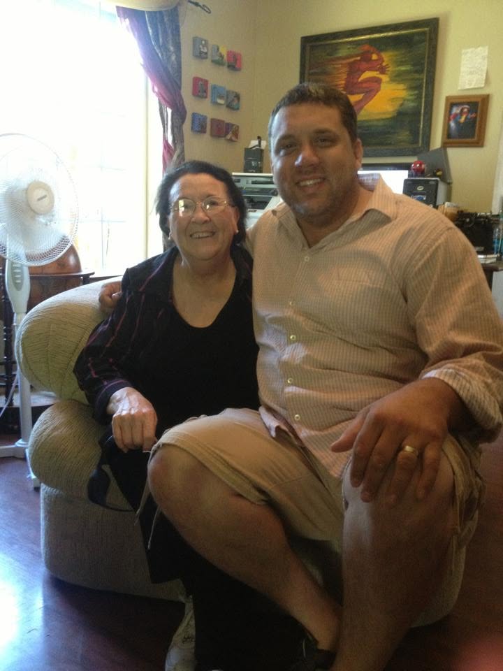 A smiling man and woman sit together on a couch in a cozy living room, surrounded by personal items and decorations.