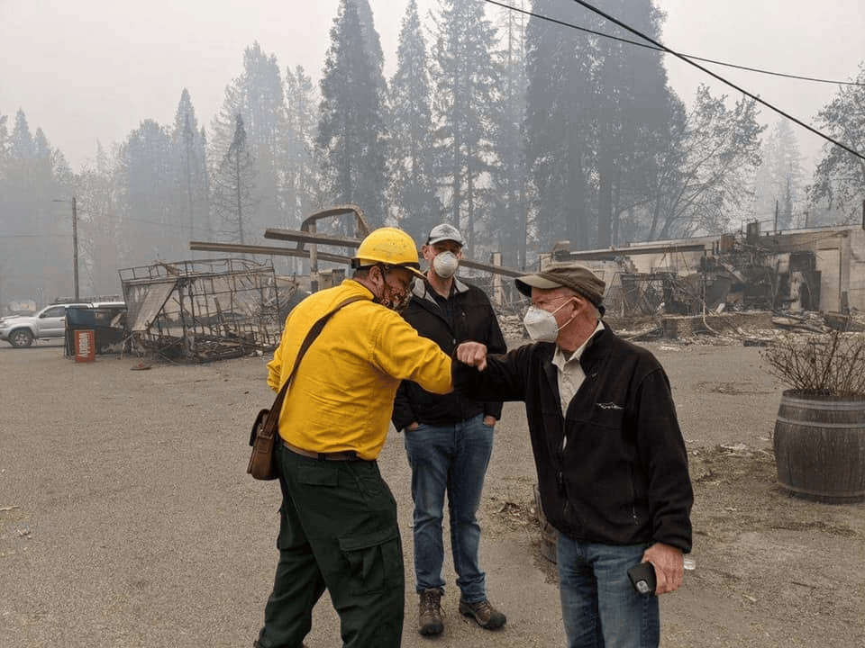Two men wearing face masks greet each other with an elbow bump, while a third man observes in a smoky, wildfire-affected area with burned structures in the background.