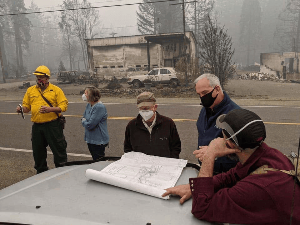 Group of people discussing plans over a blueprint in a smoke-filled area with a burnt building in the background.