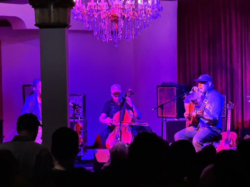 A musician playing acoustic guitar and singing on stage, accompanied by a cellist, under moody purple lighting with a chandelier in the background.