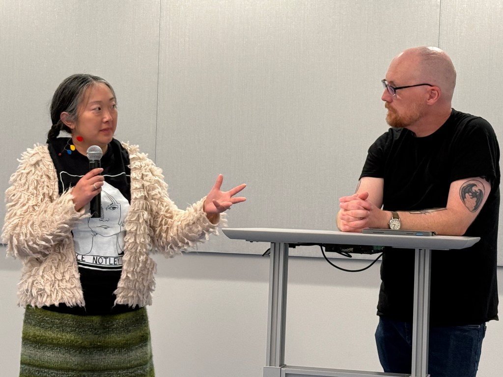 A woman with long hair and a fluffy light-colored sweater holds a microphone while speaking, gesturing with her hand. She is standing next to a man with glasses and a beard, who is seated at a table, listening attentively.
