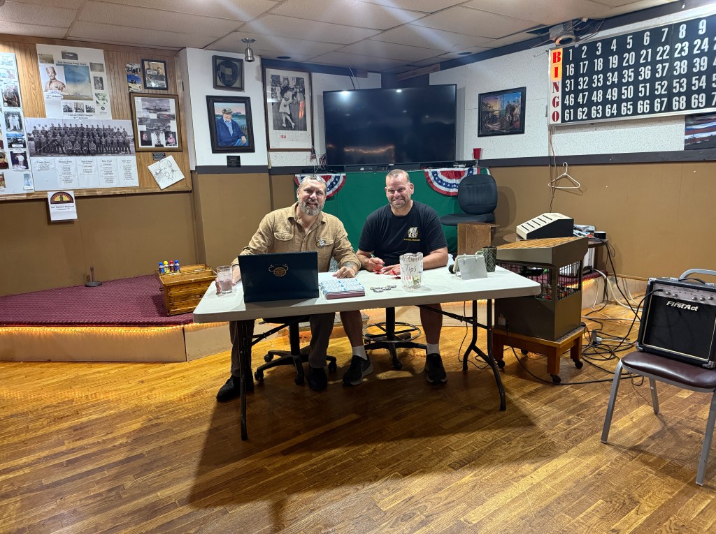 Two men sitting at a table in a recreational space, surrounded by memorabilia and decorations. One man is wearing a tan shirt and the other is in a black t-shirt. The table has cups, papers, and a tips jar. In the background, there are various framed photos and a bingo board on the wall.