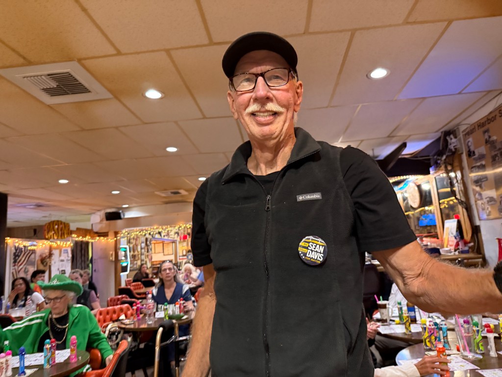 An elderly man wearing glasses and a cap stands in a lively restaurant setting, smiling at the camera. He has a button on his jacket with the name 'SEAN DAVIS' displayed. In the background, patrons are seated at tables decorated with colorful drinks.