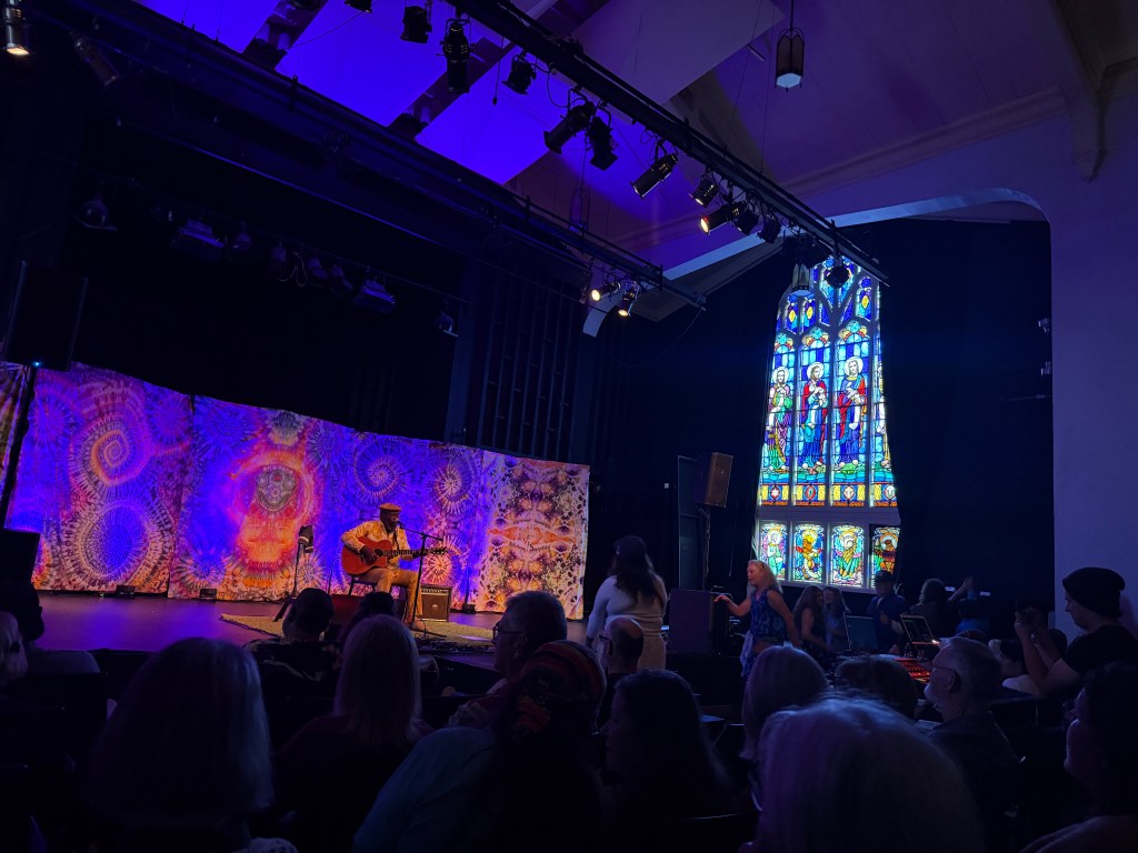 A musician playing guitar on stage in front of a colorful backdrop with abstract patterns, illuminated by purple lights, with an audience seated in front, and a stained glass window visible to the right.