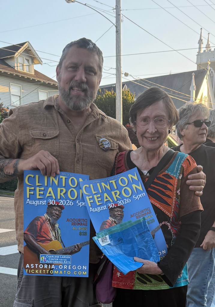 A man and a woman stand together, both smiling and holding posters for a Clinton Fearon concert on August 30, 2026. The man has a beard and wears a button on his shirt, while the woman has glasses and a colorful top. They are outdoors with buildings and trees in the background.