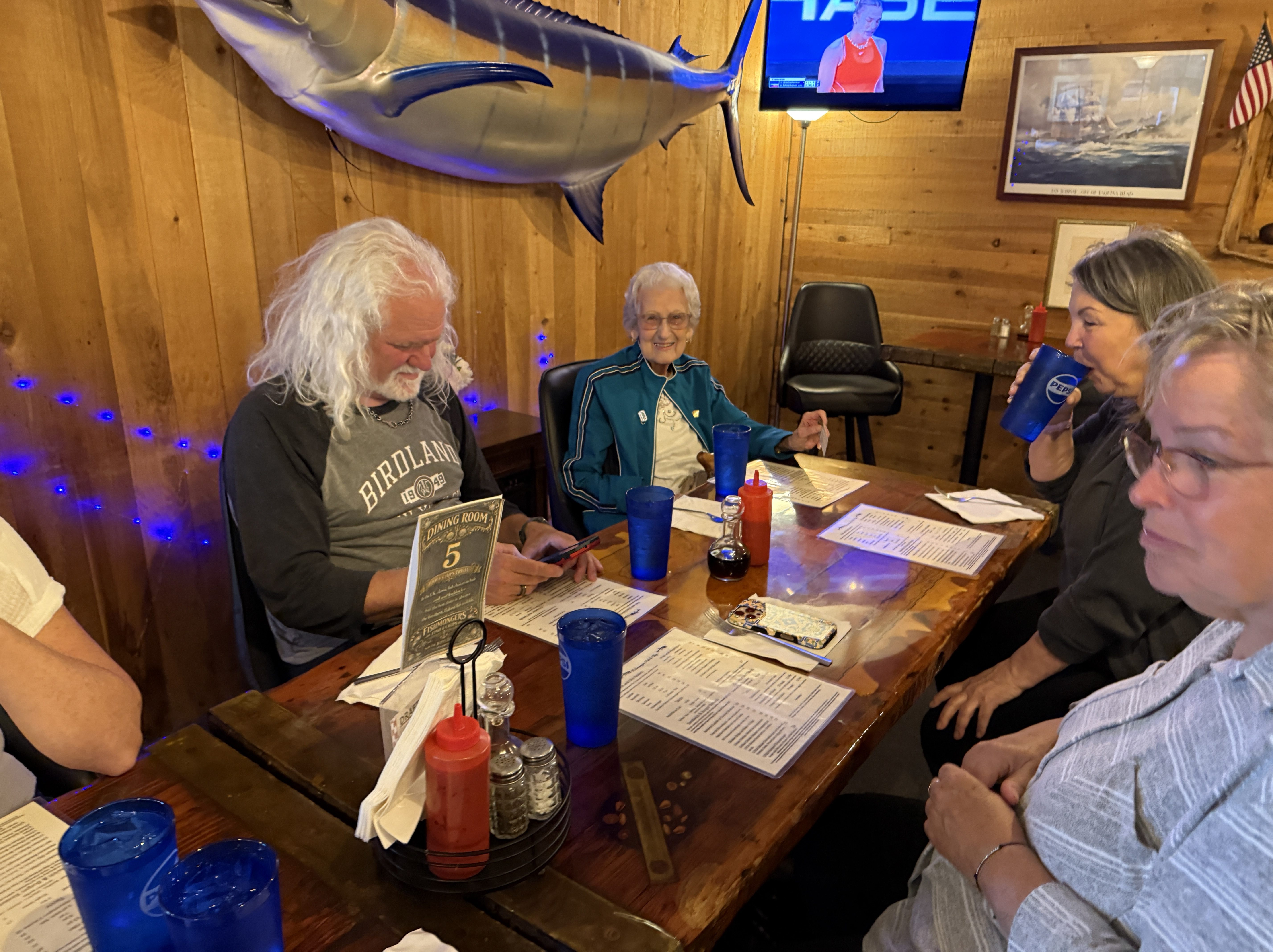 A group of four people seated at a restaurant table, reviewing menus. The setting features wooden walls and a large fish decoration above. One person has long white hair, while another is an elderly woman in a teal jacket. There are blue drinking glasses, along with condiments and a menu on the table.