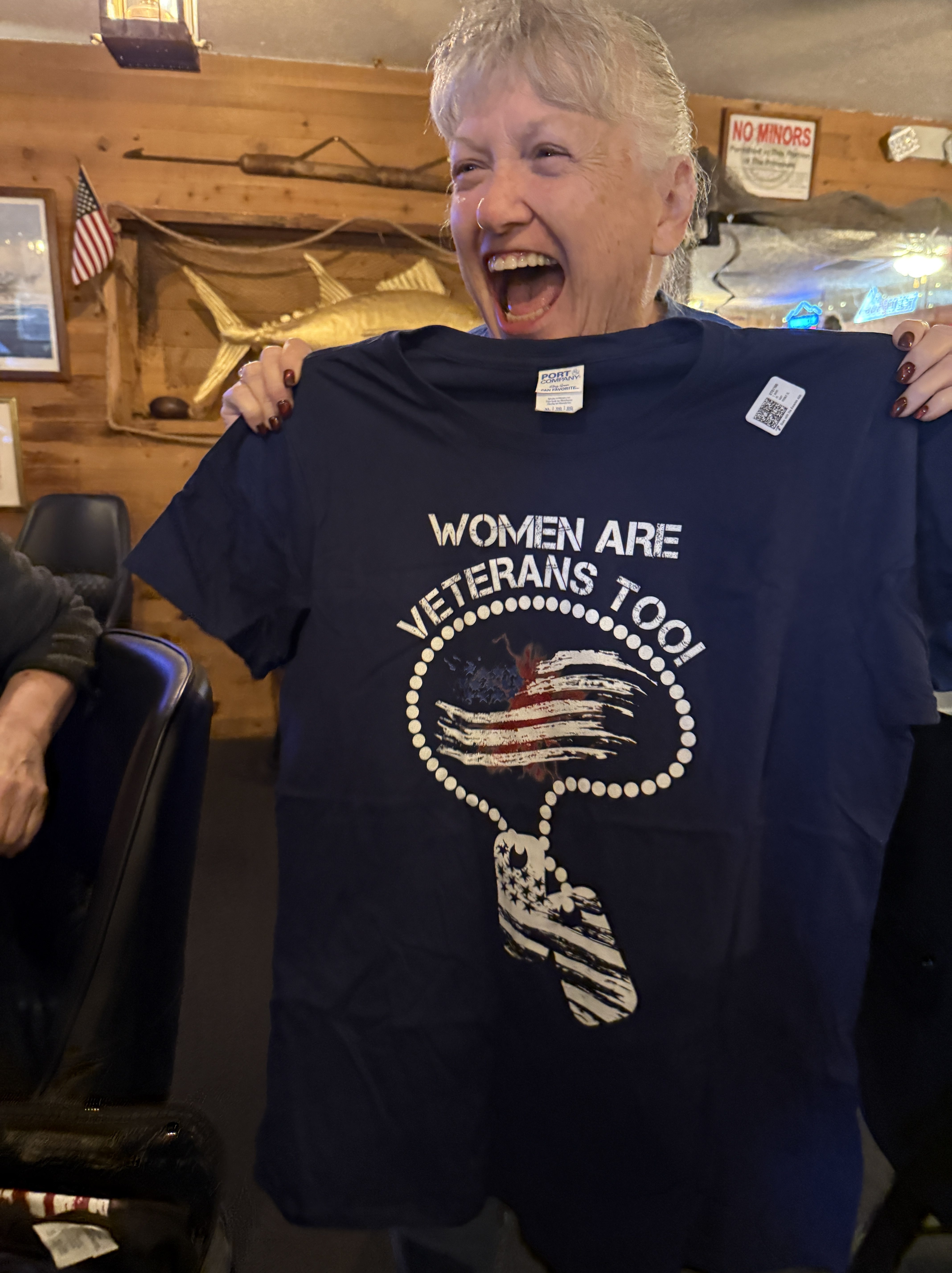 A woman joyfully holding a navy blue t-shirt that reads 'WOMEN ARE VETERANS TOO!' with patriotic designs, smiling in a lively setting.