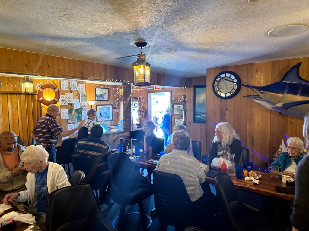 A cozy restaurant interior with a mix of customers, including older adults engaged in conversation, surrounded by nautical-themed decor, including a large fish mounted on the wall and various pictures and drawings. The space has wooden paneling and warm lighting, creating a welcoming atmosphere.