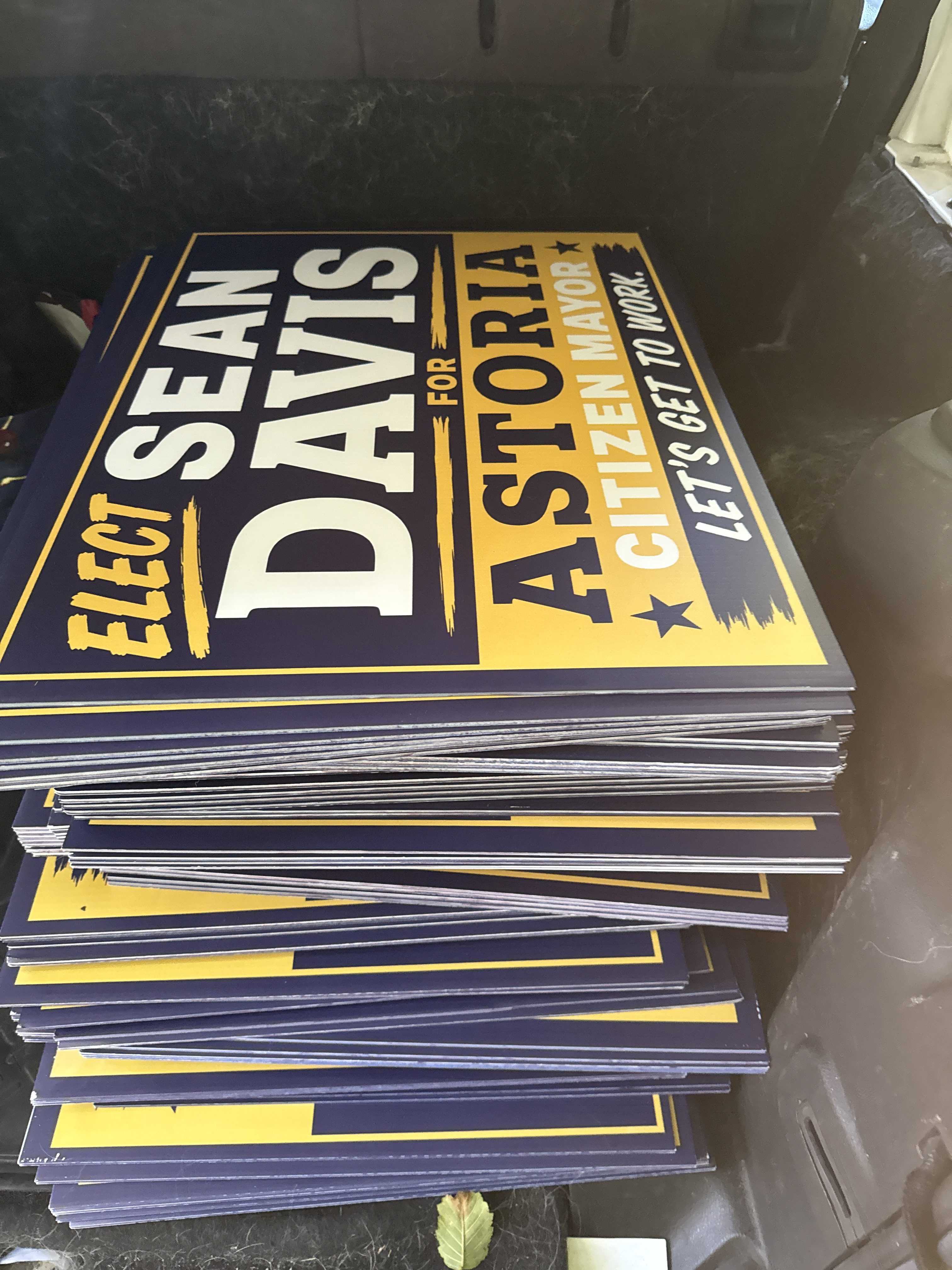 Stack of campaign signs for Sean Davis, running for Citizen Mayor of Astoria, featuring bold text and a yellow and blue color scheme.