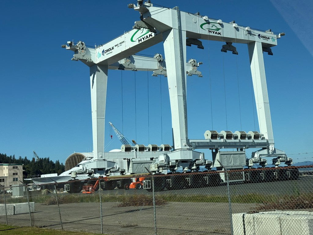 A large mobile boat hoist at a marina, with a clear blue sky in the background. The hoist features a white structure with multiple wheels and is positioned near docked boats.