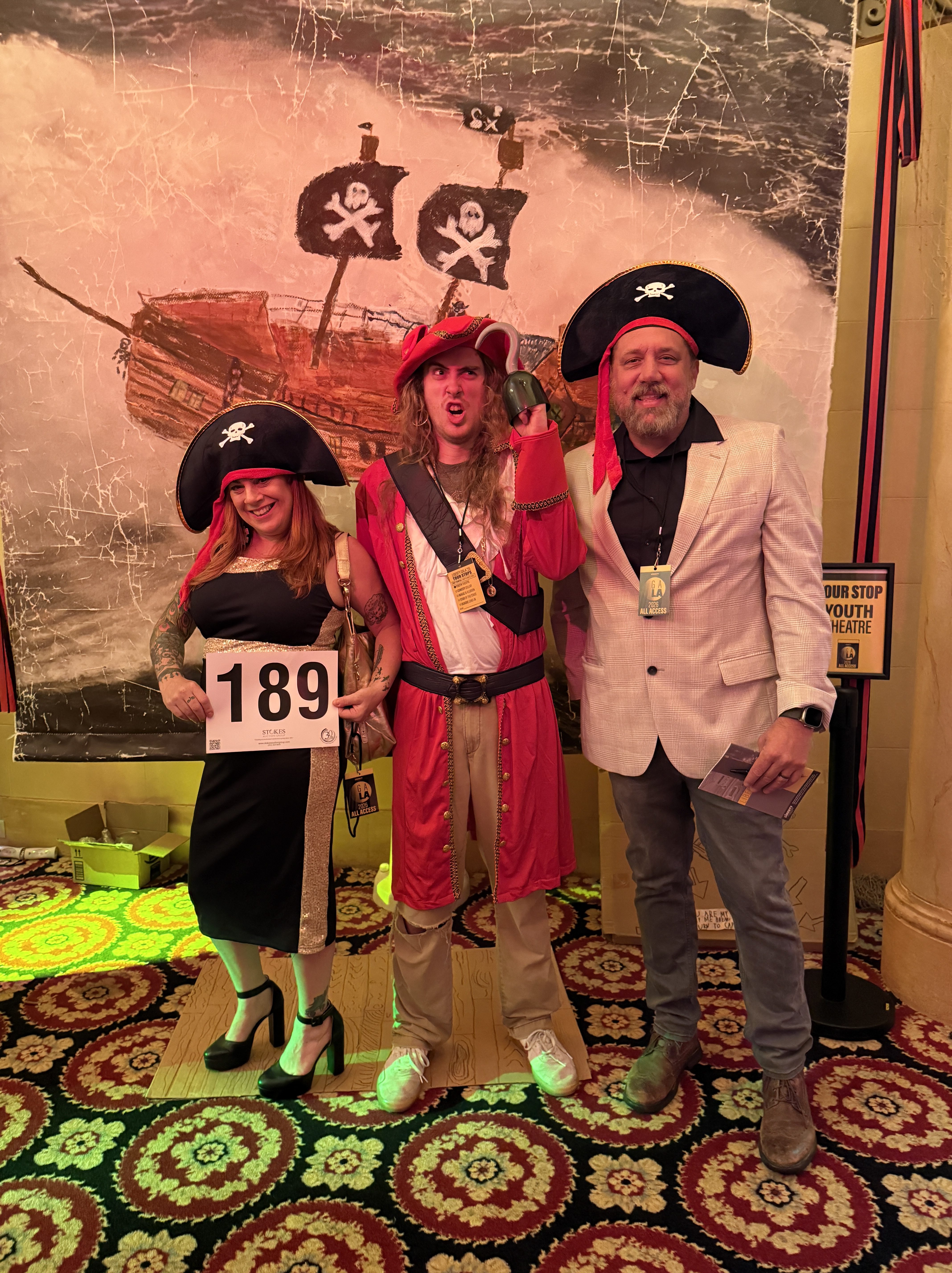 Three individuals dressed in pirate costumes posing together in front of a pirate-themed backdrop featuring a ship with skull and crossbones flags.