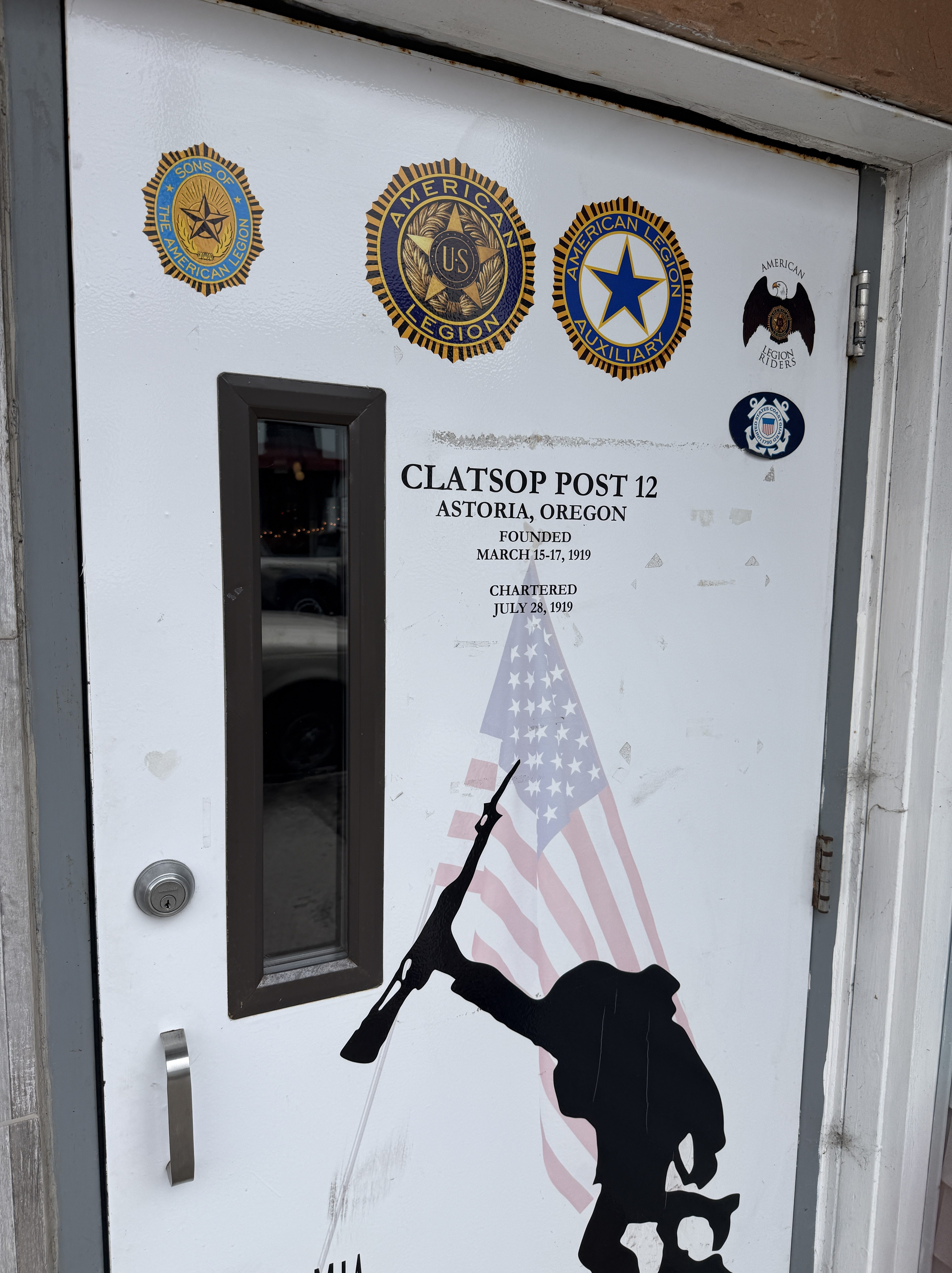 Door of Clatsop Post 12 displaying emblems of the American Legion and its affiliates, alongside details of its foundation and charter dates, set against a backdrop featuring an American flag and a silhouetted soldier.