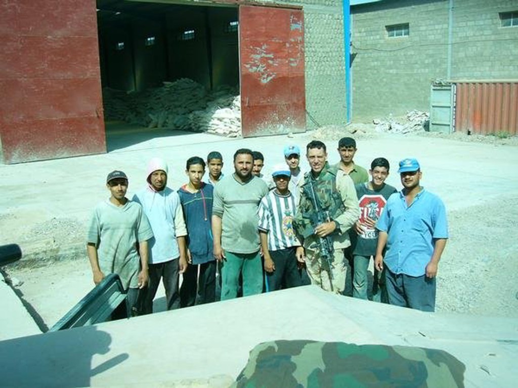 A group of men, including a soldier, posing together outdoors in front of a warehouse. The soldier is wearing camouflage and holding a firearm, surrounded by local men of various ages, standing on a gravel surface.