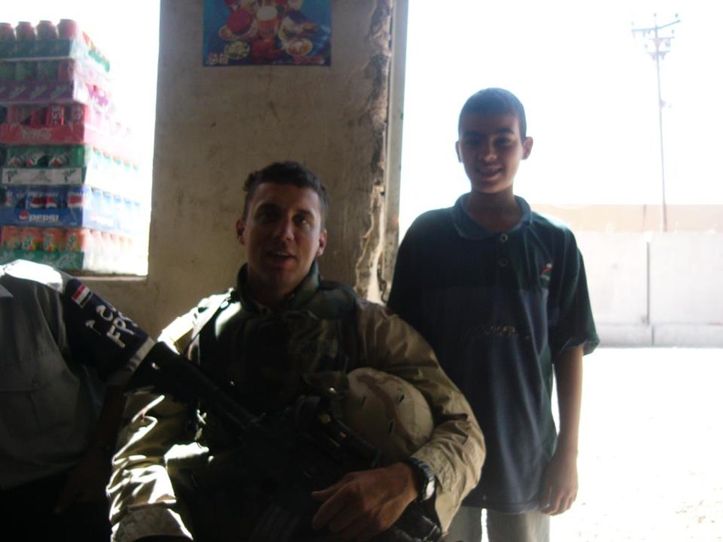 A soldier in military attire sitting against a wall with an armed rifle, next to a young boy smiling; a backdrop of colorful soda cans is visible.