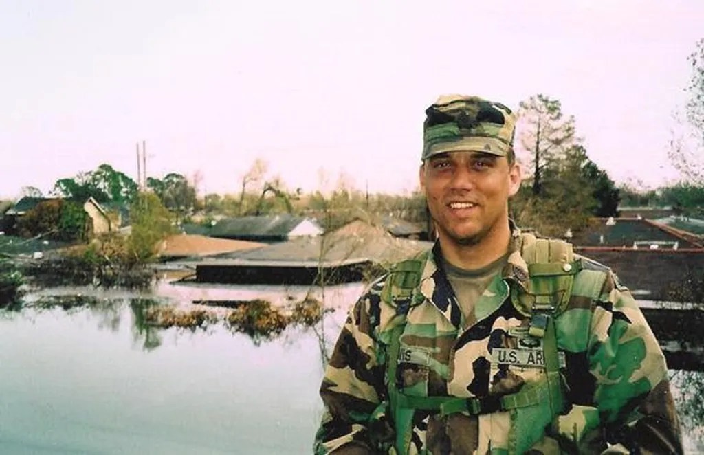 Smiling soldier in camouflage uniform standing near floodwaters, with submerged houses and trees in the background.