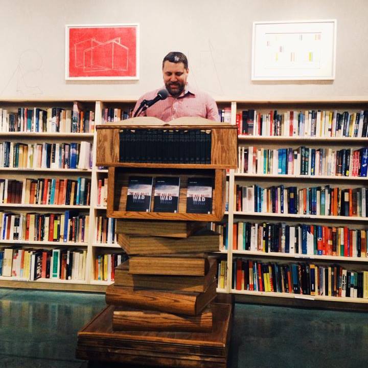 A man in a pink shirt stands at a wooden podium made of stacked books, reading from an open book. Behind him is a large bookshelf filled with various books, and framed artworks are displayed on the wall.