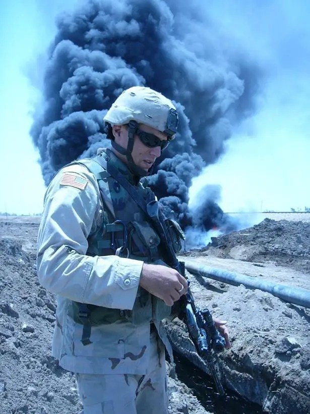 A soldier in military gear, holding a weapon, stands in front of a large plume of black smoke rising from a distant fire.