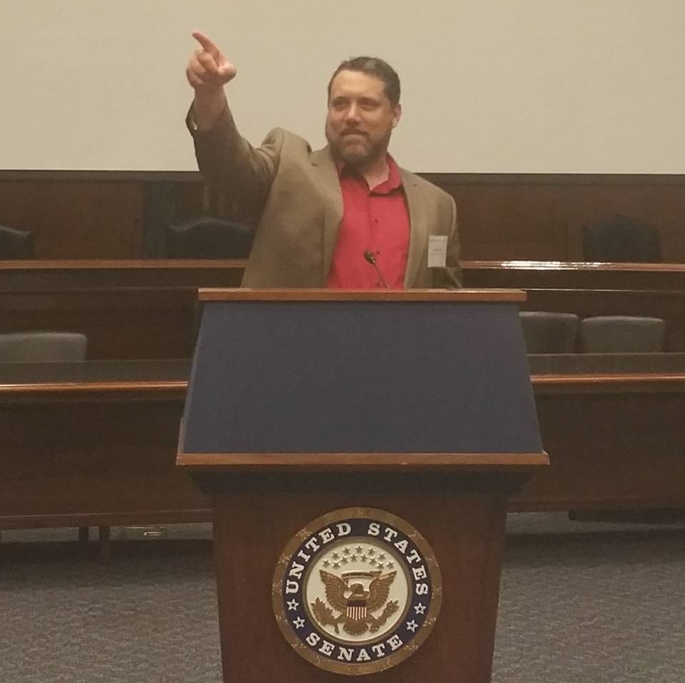 A man in a blazer and red shirt gestures while standing at a podium with the United States Senate emblem in a formal setting.