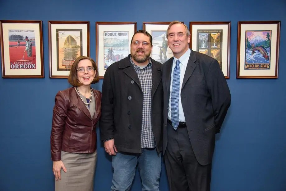 Three individuals posing for a photo in front of a blue wall decorated with framed posters of Oregon landscapes.