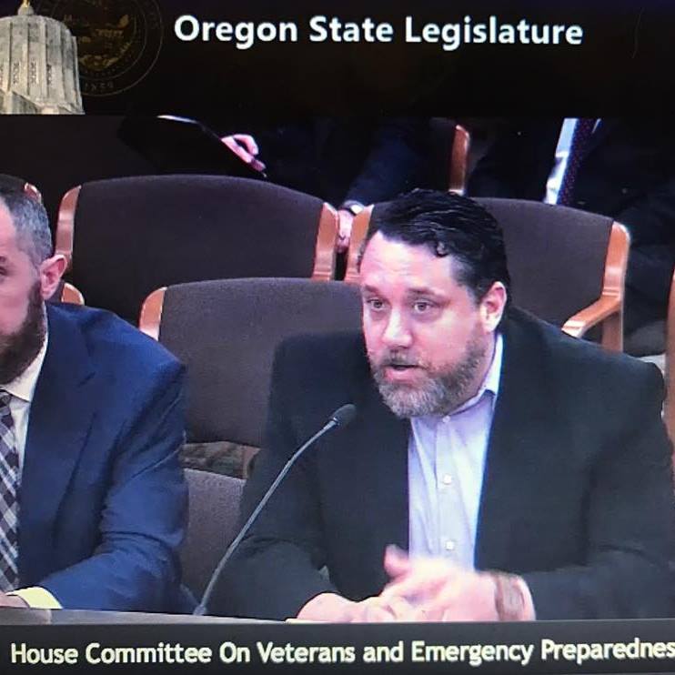 A man with a beard speaking into a microphone at a committee meeting in the Oregon State Legislature, with another man seated next to him.